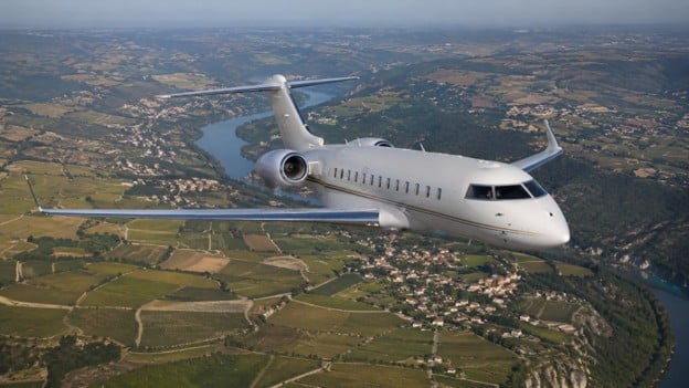 Luxury Global 6000 private jet flying over the French countryside with vineyards, villages, and a winding river below.